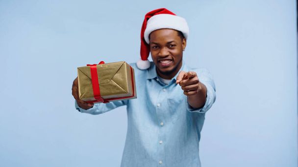 サンタ・ハットの幸せなアフリカ系アメリカ人男性が青い上に孤立したカメラを指差しながらプレゼントを包み  - 写真・画像
