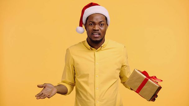 confused african american man in santa hat holding present and showing shrug gesture isolated on yellow  - Photo, Image