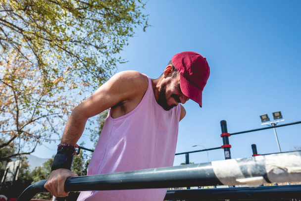 Concentrated, bearded, healthy and fit young latin man with purple shirt and cap doing a calisthenic exercise routine (performs dips on parallel bars) in an street workout park in sunny day - Photo, Image
