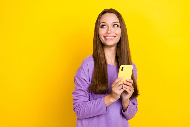 Foto van dromerig charmant meisje dragen stijlvolle kleding paars shirt houden telefoon dromerig positief uitziende lege ruimte nieuwe app geïsoleerd op gele kleur achtergrond. - Foto, afbeelding