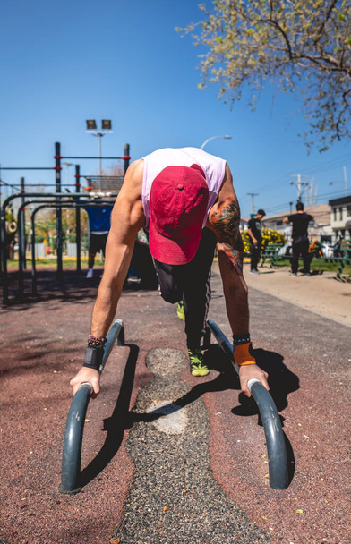 Zdravý a fit mladý latinský muž s vytetovaným ramenem a paží, čepicí a purpurovou košilí, který dělá kalistheniku (pomocí paralel) v pouličním tréninkovém parku za slunečného dne - Fotografie, Obrázek