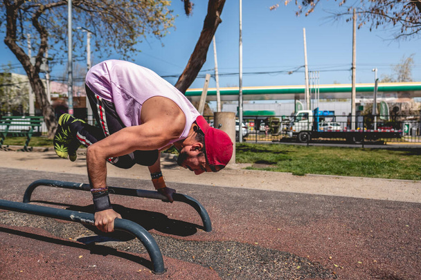 Homem latino jovem saudável e apto com braço e ombro tatuados, boné e camisa roxa fazendo calistenia (usando paralelos) em um parque de treino de rua em um dia ensolarado - Foto, Imagem