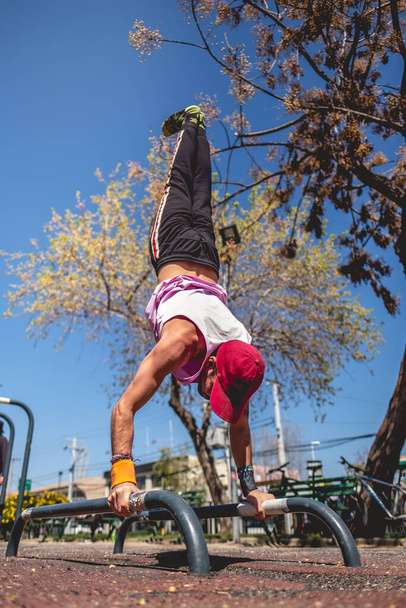 Homem latino jovem saudável e apto com braço e ombro tatuados, boné e camisa roxa fazendo calistenia (pose invertida) em um parque de treino de rua em um dia ensolarado - Foto, Imagem
