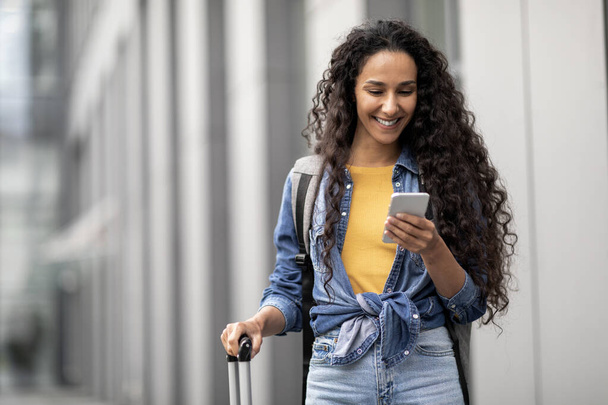 Happy attractive middle eastern young lady in casual travelling alone, carrying backpack and luggage, using smartphone while walking by airport, making online check-in, copy space - Foto, Bild