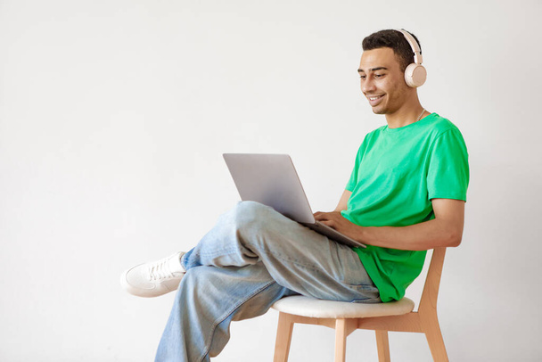 Cheerful arab man working on laptop and wearing headphones, sitting on chair over light studio wall, copy space. Guy using computer, enjoying distance job opportunities - Photo, Image
