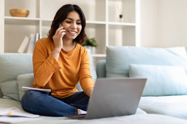 Zdalny biznes. Młoda kobieta z Bliskiego Wschodu korzystająca z telefonu komórkowego i laptopa w domu, uśmiechnięta arabska freelancer pracująca z komputerem i rozmawiająca przez telefon komórkowy siedząc na kanapie w salonie - Zdjęcie, obraz