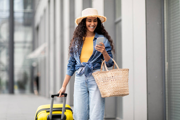 Joyeux jolie jeune femme bouclée dans une tenue décontractée confortable et chapeau de paille aller vacances seul, marcher par l'aéroport avec des bagages jaunes, en utilisant un smartphone, réservation d'hôtel en ligne, espace de copie - Photo, image