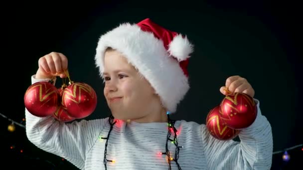 Een gelukkige 5-jarige jongen in een kerstmuts heeft plezier met rode kerstballen - Video