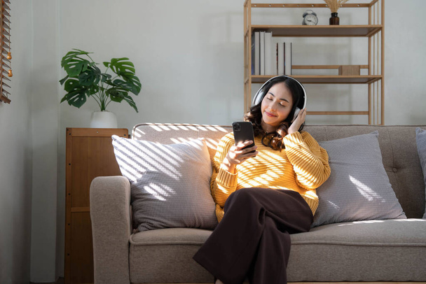 Χαμογελαστή νεαρή γυναίκα κάθεται στον καναπέ στο σπίτι μιλάμε για βιντεοκλήση στο smartphone με ακουστικά. Ευτυχισμένη χιλιετή γυναίκα στα ακουστικά χρησιμοποιούν το κινητό τηλέφωνο έχουν κάμερα ψηφιακή online επικοινωνία.. - Φωτογραφία, εικόνα