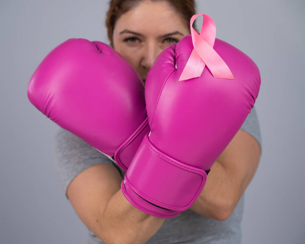 Mujer caucásica con guantes de boxeo rosa con una cinta rosa en el pecho sobre un fondo gris. Lucha contra el cáncer de mama - Foto, Imagen
