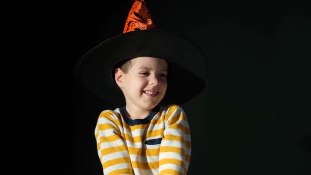 Счастливый 5-летний мальчик на Хэллоуин в шляпе ведьмы и полосатой оранжевой куртке на черном фоне. - Кадры, видео