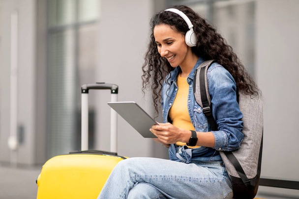 Позитивная молодая брюнетка с рюкзаком и чемоданом турист с помощью цифровых планшетных и беспроводных наушников, сидя на скамейке на открытом воздухе, смотреть фильм онлайн. Развлечения во время путешествия - Фото, изображение