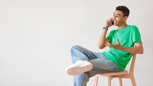 Pomysły na biznes, nowoczesną pracę i komunikację. Młody arabski mężczyzna rozmawiający przez telefon komórkowy i gestykulujący, siedzący na krześle odizolowany na tle ściany studia światła, panorana z pustą przestrzenią - Zdjęcie, obraz