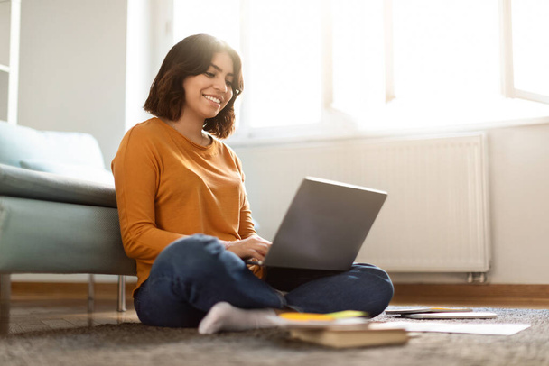 Дистанционное обучение. Молодая арабская студентка с ноутбуком дома, улыбающаяся женщина из Ближнего Востока, сидящая на полу и печатающая на компьютере, наслаждающаяся онлайн-образованием, копирующая пространство - Фото, изображение