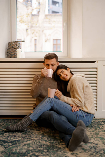 Romantikus pár otthon. Egy vonzó fiatal nő és jóképű férfi élvezik az együtt töltött időt, miközben egy csésze teával a kezükben babusgatják egymást.. - Fotó, kép
