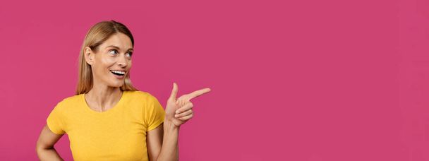 Smiling adult european woman with open mouth pointing finger at empty space, isolated on purple background, studio, close up, panorama. Choice, shocking offer and ad, huge sale and people emotions - Фото, изображение