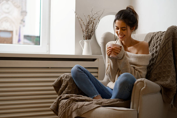 Smiling lady in smart trendy wear is sitting on armchair with a cup of tea. Smiling, sitting in relaxing atmosphere indoors at home, nice design. - Photo, Image