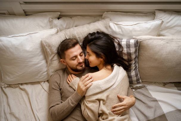 Όμορφο νεαρό ευτυχισμένο ζευγάρι που χαλαρώνει στο κρεβάτι και χαμογελά, αγκαλιάζει - Φωτογραφία, εικόνα