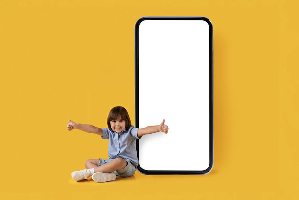 Content for kids. Excited little boy sitting near big smartphone with blank white screen and gesturing thumbs up, smiling to camera, empty space - Fotografie, Obrázek