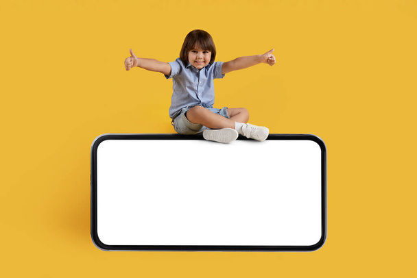 Cheerful little boy sitting on big mobile phone with empty screen and gesturing thumbs up, smartphone in landscape position with free space, orange studio background - 写真・画像