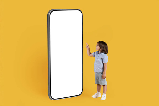 Захватывающие современные технологии. Очаровательный маленький мальчик указывает на большой пустой смартфон с белым экраном, выбирая контент на устройстве, оранжевый фон студии, свободное место - Фото, изображение