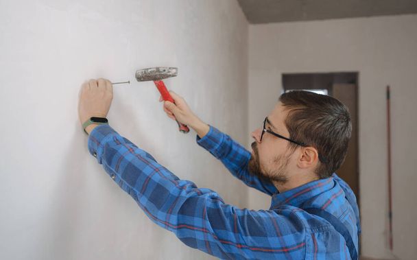 νεαρός άνδρας με μπλε κοστούμι κάνει επισκευή διαμέρισμα. Σχέδιο ανακαίνισης σπιτιού. Ευτυχισμένος χαμογελαστός μάστορας σφυρηλατεί καρφιά στον τοίχο - Φωτογραφία, εικόνα