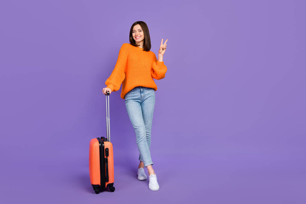 Full size photo of pretty young girl show v-sign baggage business trip wear trendy orange knitwear look isolated on violet color background. - Фото, зображення