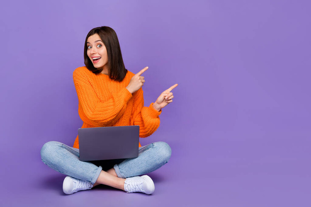 Full length photo of nice young lady point excited empty space laptop wear trendy orange knitwear look isolated on violet color background. - Zdjęcie, obraz