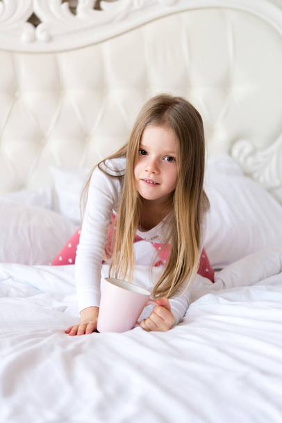 Little cute girl has breakfast in bed.  girl is holding a cup. light interior. - Foto, Imagen