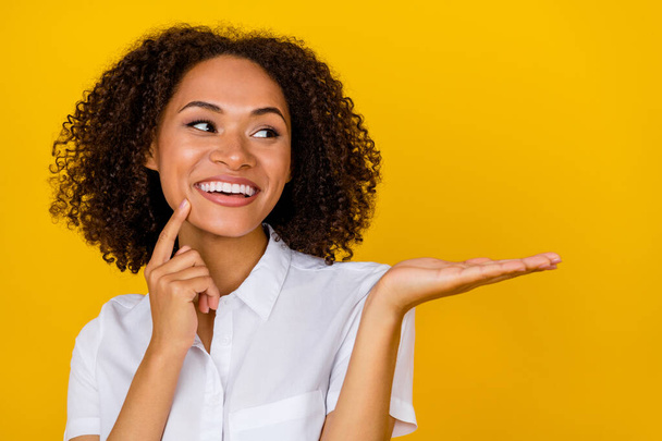 Photo of cheerful positive toothy girl wavy hairdo finger on cheekbone chin palm directing empty space isolated on yellow color background. - 写真・画像