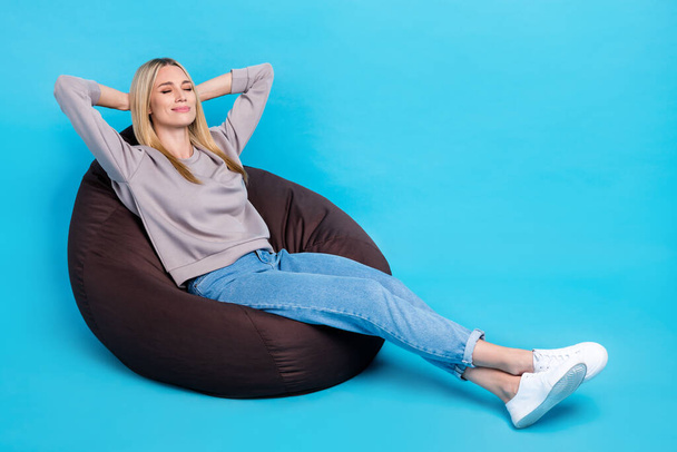 Full length photo of adorable cute girl dressed grey sweatshirt sitting bean bag arms behind head empty space isolated blue color background. - Фото, изображение