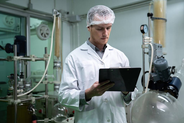 Apothecary scientist using a clipboard and pen to record information from a CBD oil extractor and a scientific machine used to create medicinal cannabis products. - Foto, Imagen