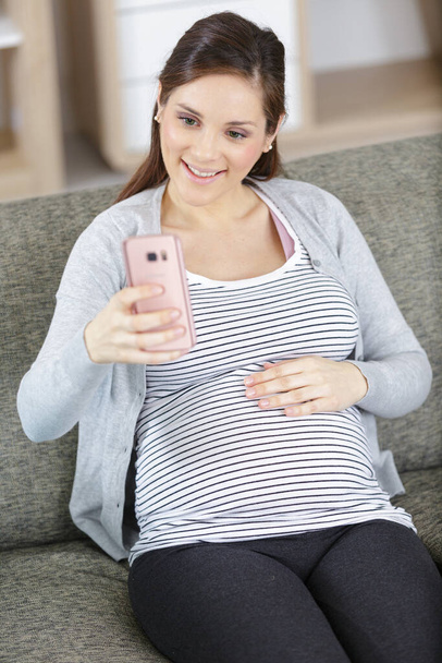 pregnant woman sitting on sofa and taking selfie - Zdjęcie, obraz