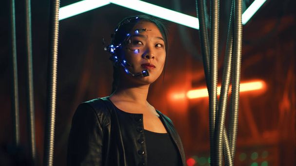 Cyberpunk girl in a black leather jacket and intense facial expressions. Asian girl with one-eyed glasses and headset. Trapped behind black hanging rods. Cyborg, sci-fi, Neon lights. - Φωτογραφία, εικόνα