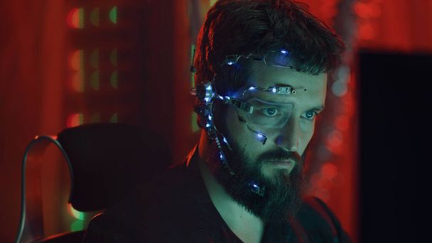 Un tipo Cyberpunk mira la pantalla de la computadora. Usando gafas futuristas de un solo ojo con auricular y micrófono. Fondos cibernéticos y de ciencia ficción. - Foto, imagen