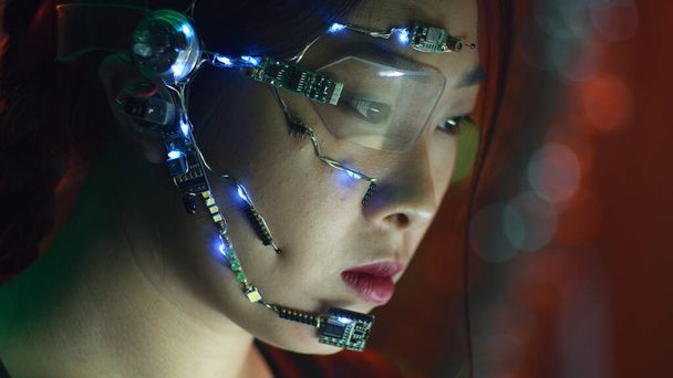 Focalisé fille asiatique en tenue cyberpunk fonctionne sur l'ordinateur. Porter des lunettes à un œil avec des lumières LED blanches et un microphone. Fond néon lumières. science fiction, concept de cybernétique. - Photo, image