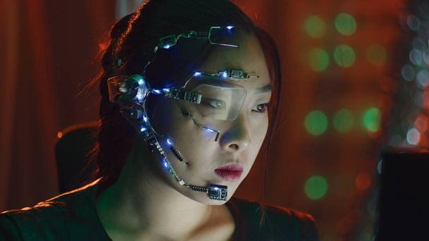 Μια Cyberpunk δουλεύει στον υπολογιστή με τα κόκκινα φώτα νέον. Ασιάτισσα με φουτουριστικό μονόφθαλμο γυαλιά και μικρόφωνο. Διαδικτυακό υπόβαθρο και υπόβαθρο επιστημονικής φαντασίας. - Φωτογραφία, εικόνα