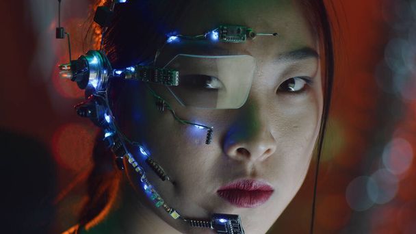 Una chica Cyberpunk con auriculares con micrófono, gafas de un ojo y pequeñas luces led blancas. Luces rojas de neón en el fondo. Chica asiática vestida de cyberpunk. Fondos cibernéticos y de ciencia ficción. - Foto, Imagen