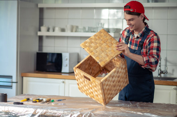 Χαμογελώντας ευχαριστημένος επικεντρώθηκε νεαρός ξυλουργός σε workwear προσάρτηση του αρθρωτό καπάκι στο κουτί άχυρο - Φωτογραφία, εικόνα