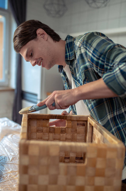 Piacevole artigiano appoggiato sul tavolo mentre spazzola la vernice sul bordo della scatola di paglia - Foto, immagini