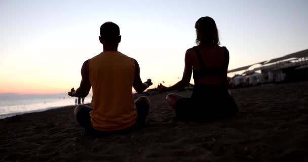 Young couple sits on seashore in lotus position at sunset and practices yoga. Family couple relax yoga and sea - Footage, Video