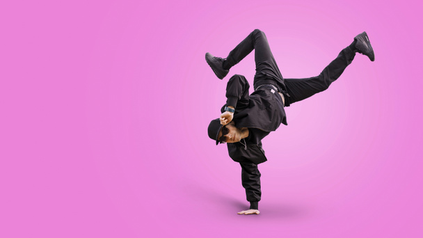 Fashion handsome man dancer with stylish baseball cap and sunglasses in a fashionable black outfit with a bomber jacket, jeans and sneakers performs on a pink background and stands on hand. Dance boy - Photo, Image