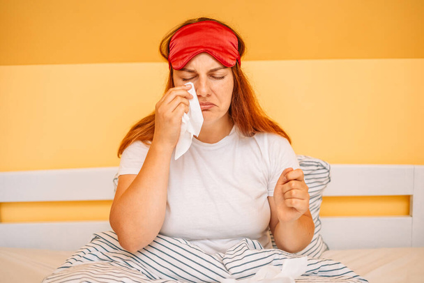 Una joven infeliz está llorando y sosteniendo un pañuelo para la nariz mientras está sentada en la cama, sintiéndose deprimida, estresada y con migraña. - Foto, Imagen