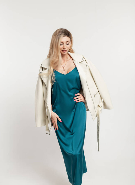 Modelo de mulher loira bonita na moda em um vestido turquesa elegante com uma jaqueta de couro bege elegante em um fundo branco no estúdio - Foto, Imagem
