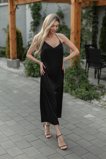 moda dość elegancki elegancki kobieta z blond włosy w stylowy czarny długo sexy strappy sukienka z buty spacery na ulicy - Zdjęcie, obraz