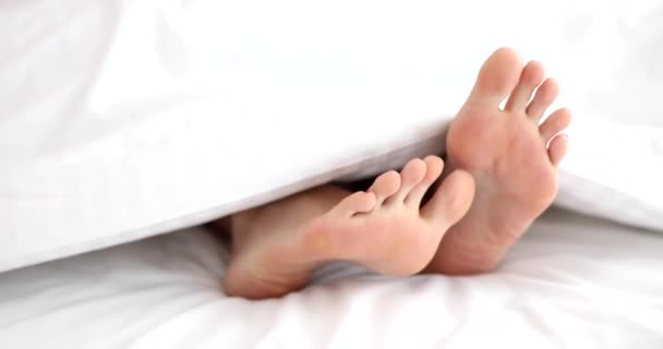 Primo piano delle gambe della donna sotto coperta bianca graffia piede. . Persona irriconoscibile che dorme a letto a casa e concetto di riposo e svago - Filmati, video