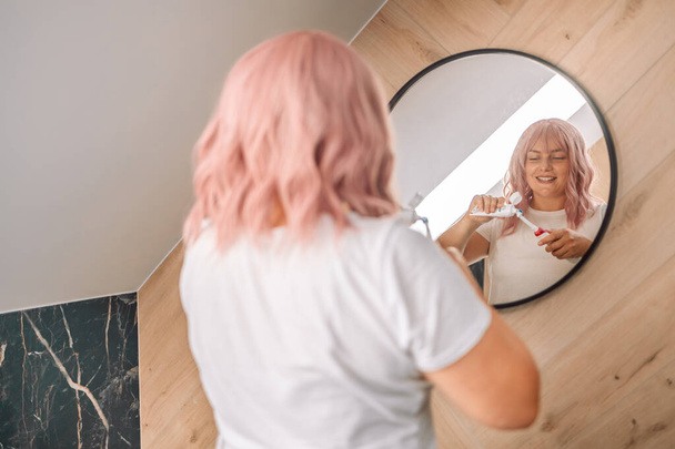 Biała różowa kobieta myjąca zęby elektryczną szczoteczką w łazience. Nowoczesny styl życia domowego. Higiena i opieka dentystyczna - Zdjęcie, obraz
