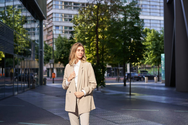 Retrato de una joven mujer corporativa confiada en traje beige, parada en la calle en su traje de negocios, con aspecto seguro de sí misma. - Foto, imagen