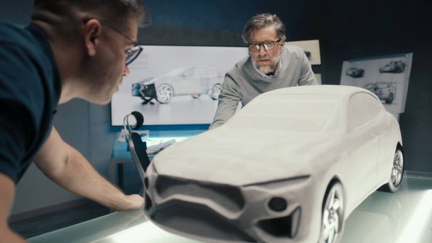 Automotive engineers work in a car design studio with a wall mounted LED. Discuss the design of the sculpture of prototype car model placed on a glass table. - Фото, зображення