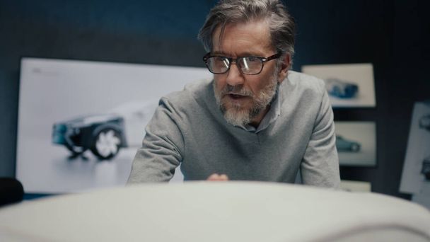 Grey haired automotive engineer in a framed glasses, discusses the design changes in the sculpture of a car with his colleague in a high tech car manufacturing company. - Photo, Image
